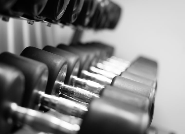 Rows of black rubber-coated dumbbells with silver handles are neatly arranged on a multi-tiered rack in a gym, captured in a black and white photograph.