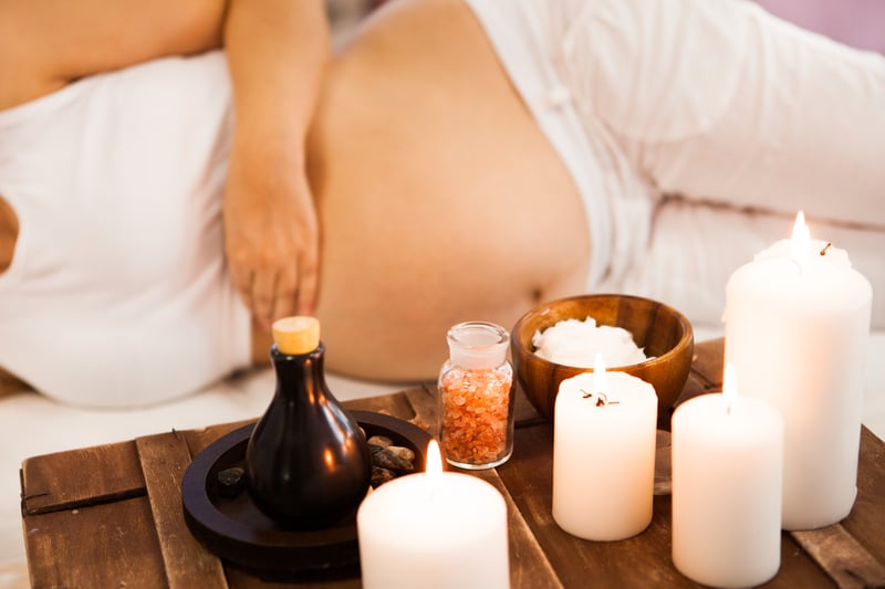 Pregnant woman lies, hand on bare belly. Lit candles, bath salts, a dark oil bottle, and cream are arranged on a wooden tray in the foreground.