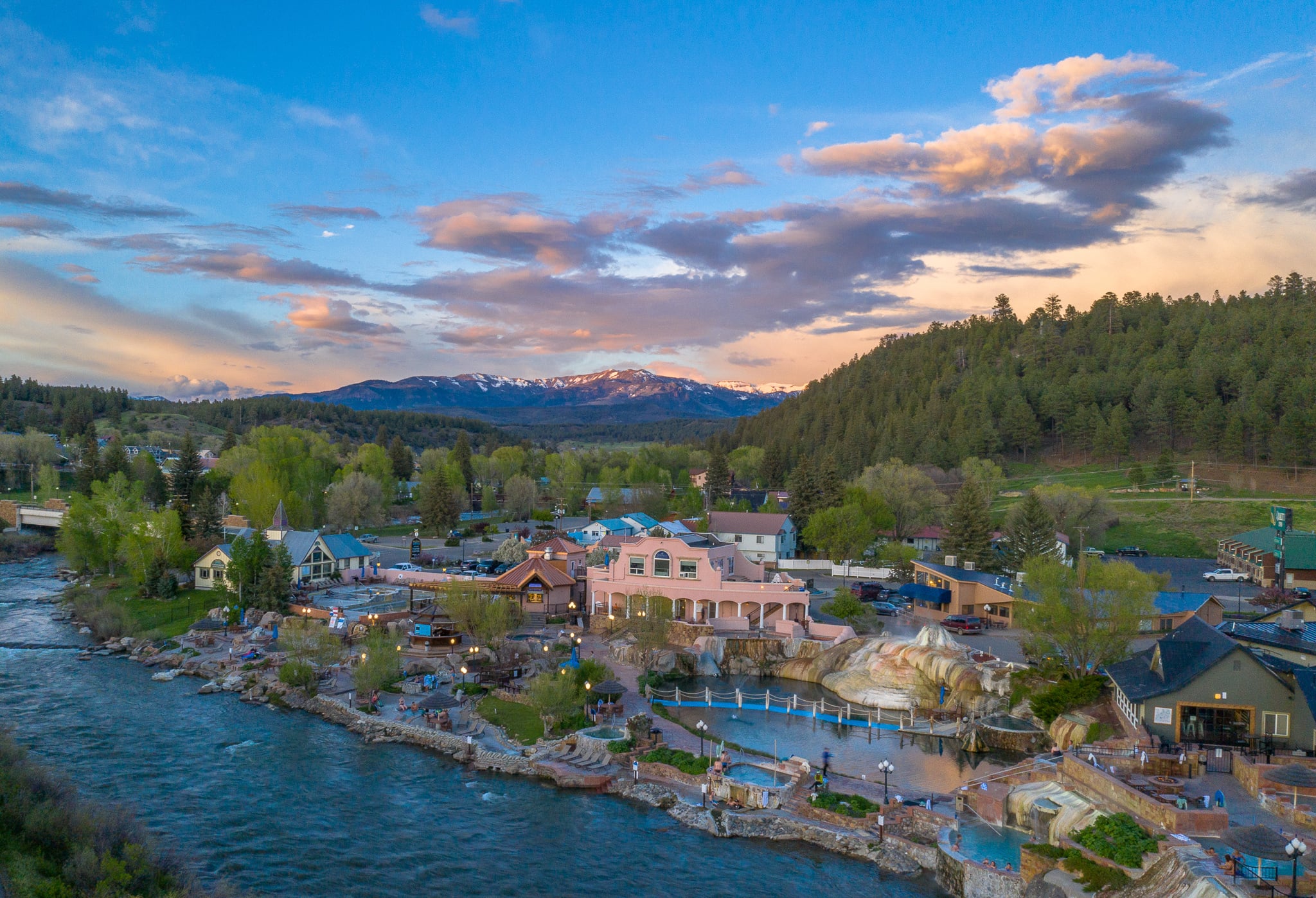 A large pink resort with multi-tiered thermal pools sits beside a winding river, within a mountain town backed by forests under a vibrant sunset sky.