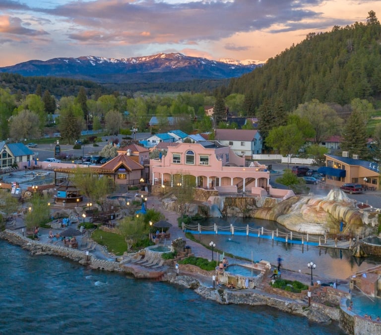 The Springs Resort (SPA) offers multiple hot springs pools along a river, surrounded by buildings, a town, and forested mountains under a colorful sunset sky.