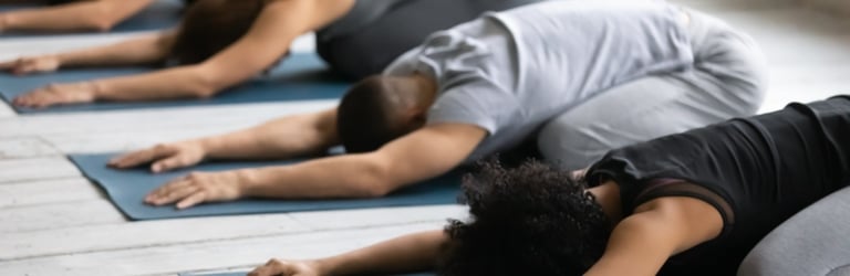 Diverse people perform Child's Pose on blue yoga mats, kneeling with outstretched arms and heads down, in a bright room with a wooden floor.