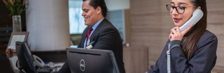 A female hotel receptionist talks on a white telephone, while a male colleague works at a computer behind a counter. DELL is visible on the monitor.