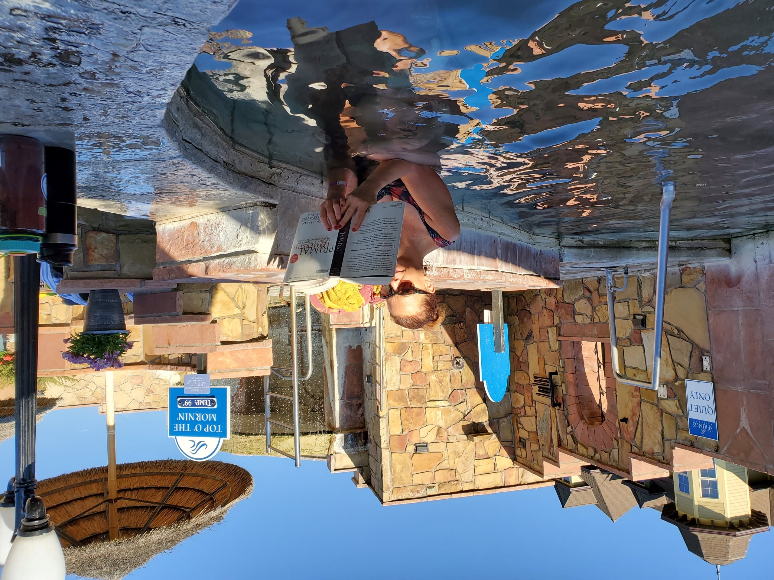 A woman wearing sunglasses reads a book while submerged chest-deep in a sunny outdoor pool with stone walls. Text: 
