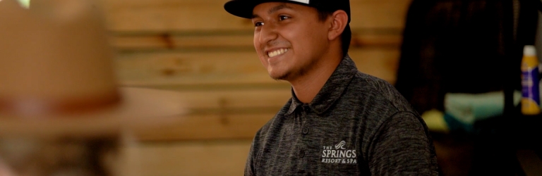 A young man smiles, wearing a black and white hat and a grey polo shirt with 