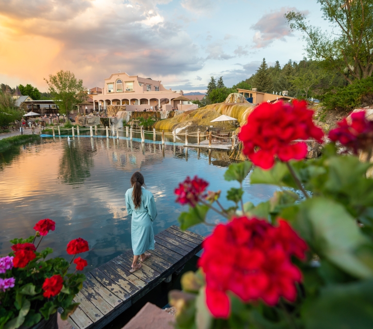 Woman in blue robe stands on a dock, facing a calm pool reflecting sunset clouds. A resort building, natural hot springs, and trees are in the background; red flowers are in the foreground.