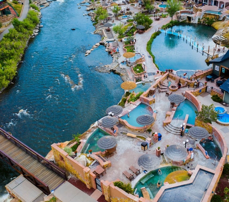 A large hot springs resort complex features numerous pools with people bathing and relaxing. It is situated alongside a wide, flowing river with a long pedestrian bridge and lush green banks.