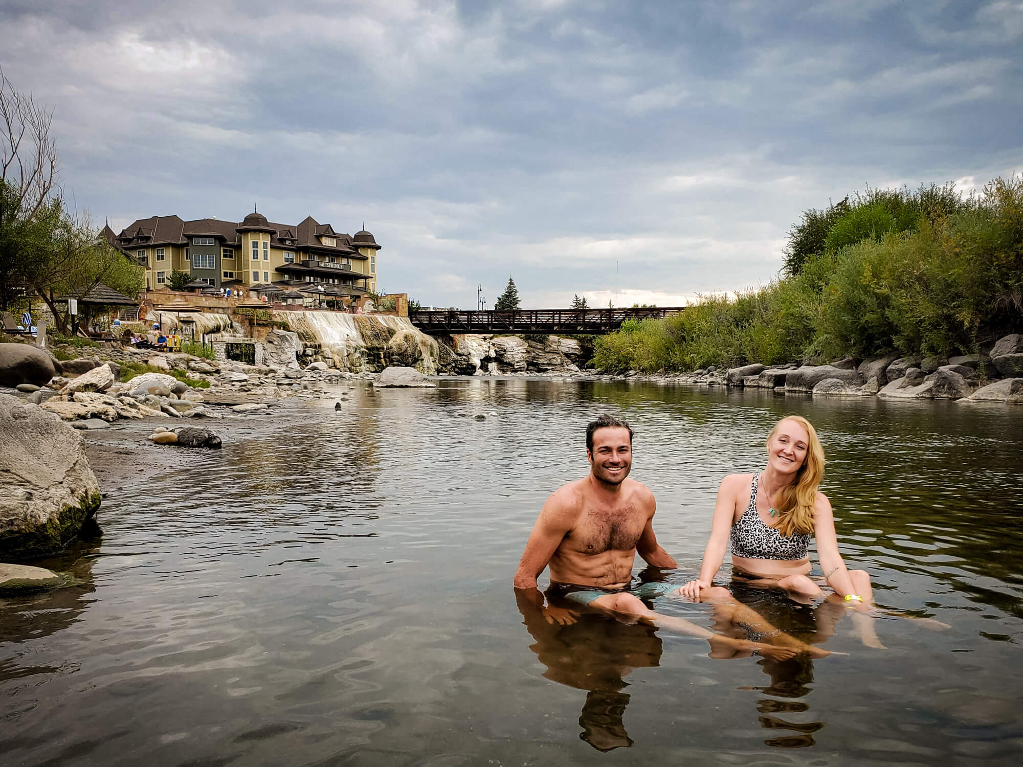 A smiling man and woman sit in a shallow river. Behind them, 