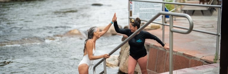 Two women high-five at the edge of a river, one standing in the water and the other on stone steps, next to a sign.