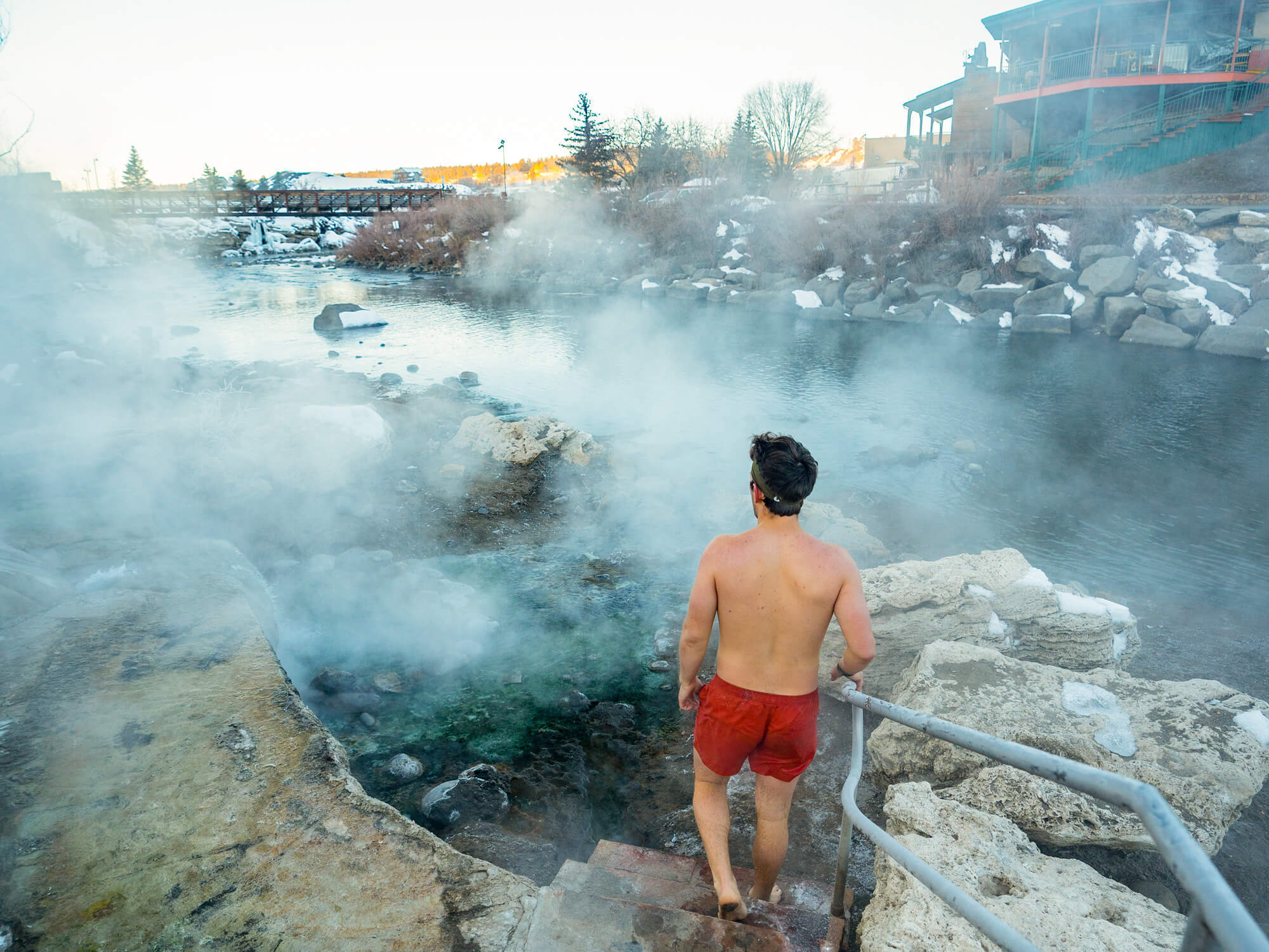A shirtless man in red shorts descends steps into a steaming natural hot spring. Snowy banks, a river, bridge, and distant buildings complete the cold-weather scene.