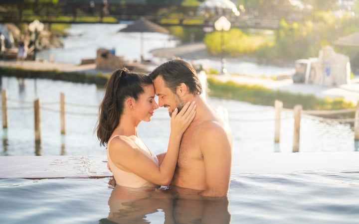 A couple, partially submerged in an infinity pool, embraces with foreheads touching, against a sunny background of a river and distant bridge.