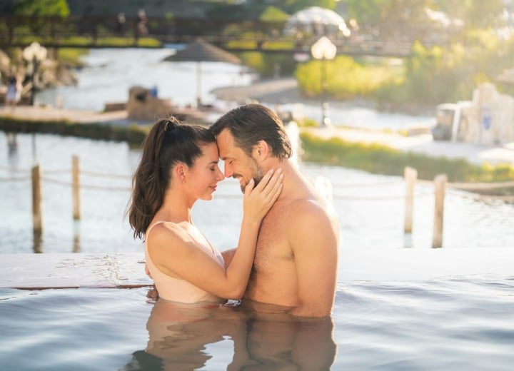 A couple, partially submerged in an infinity pool, embraces with foreheads touching, against a sunny background of a river and distant bridge.