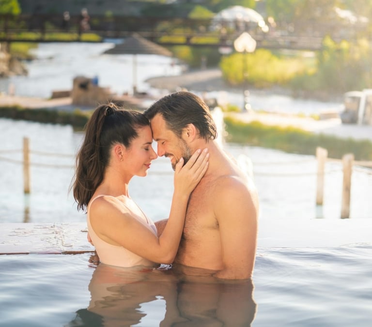 A couple, partially submerged in an infinity pool, embraces with foreheads touching, against a sunny background of a river and distant bridge.