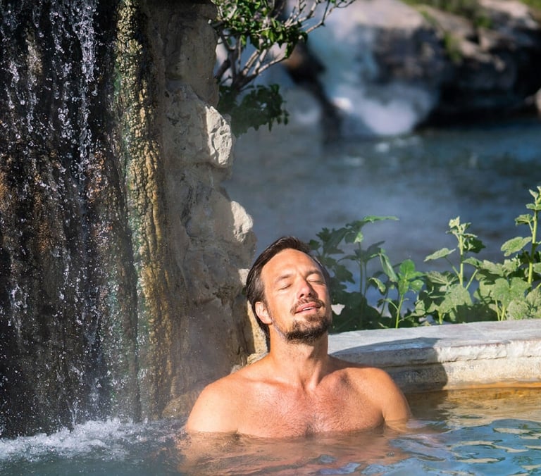 Man relaxing with eyes closed in a hot spring pool beside a rock waterfall. A sign in the background reads: HOT SPRINGS, NO DIVING, NO JUMPING, NO EATING, NO DRINKING, NO SMOKING, NO GLASS.