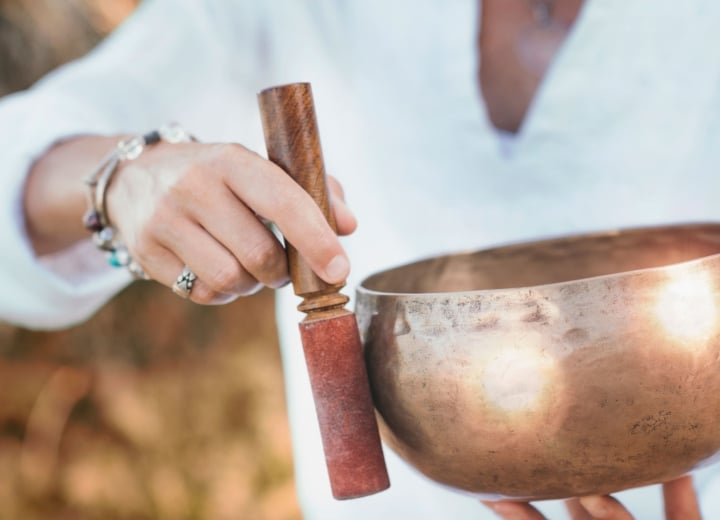 A person's hands hold a bronze singing bowl and wooden mallet, preparing to play it, dressed in white against a blurred natural background.