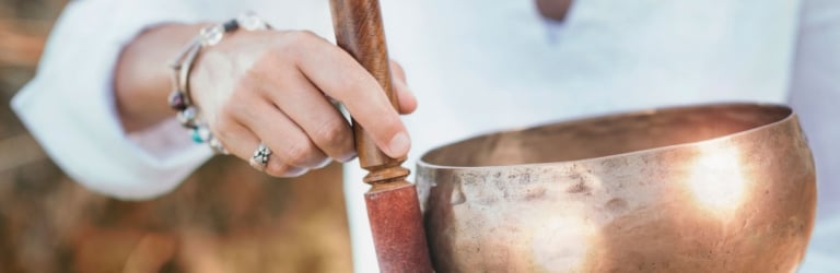 A person's hands hold a bronze singing bowl and wooden mallet, preparing to play it, dressed in white against a blurred natural background.