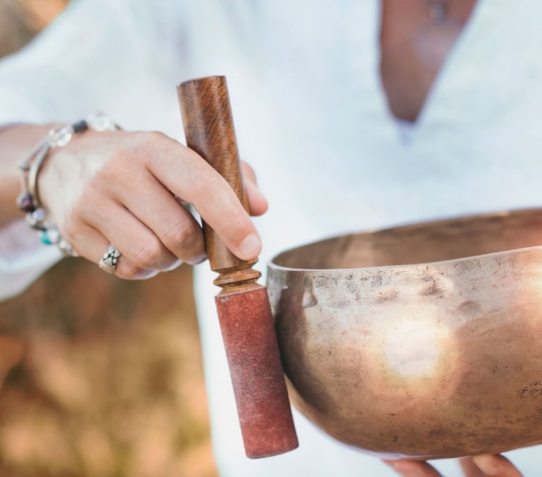A person's hands hold a bronze singing bowl and wooden mallet, preparing to play it, dressed in white against a blurred natural background.