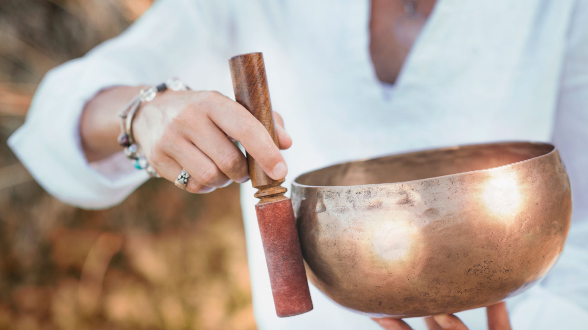 A person's hands hold a bronze singing bowl and wooden mallet, preparing to play it, dressed in white against a blurred natural background.