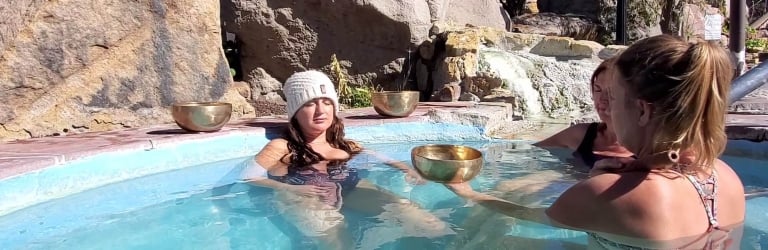 A woman in a white beanie relaxes in a light blue pool, holding a singing bowl. Another woman offers her a second bowl. Large rocks and a waterfall surround them outdoors.