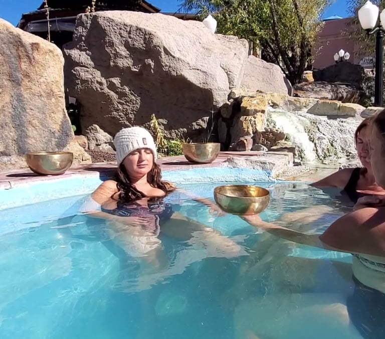 A woman in a white beanie relaxes in a light blue pool, holding a singing bowl. Another woman offers her a second bowl. Large rocks and a waterfall surround them outdoors.