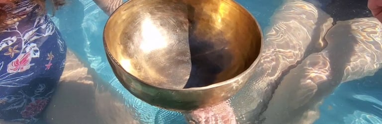 A large, golden singing bowl floats in clear blue water, supported by several people's hands and feet submerged in a pool.