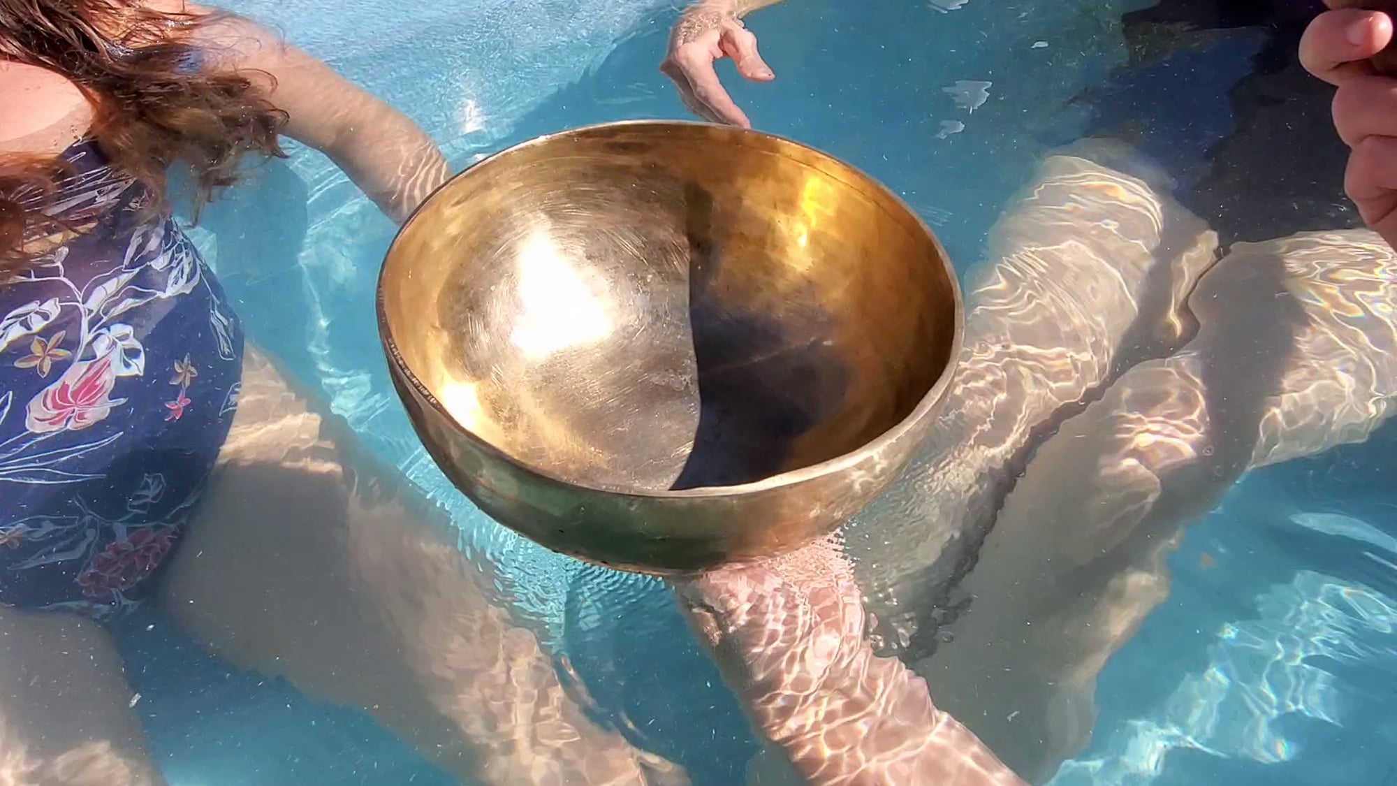 A large, golden singing bowl floats in clear blue water, supported by several people's hands and feet submerged in a pool.