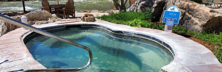 A natural-looking hot tub with clear, light green water and a handrail overlooks a rushing river, flanked by rocks and lush plants. Signs read: 