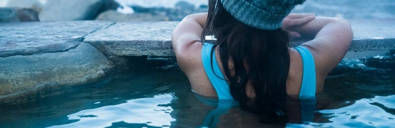 A woman wearing a blue swimsuit and gray knit pom-pom hat is submerged in a steaming outdoor pool, leaning on its stone edge. A snowy river landscape is in the background.