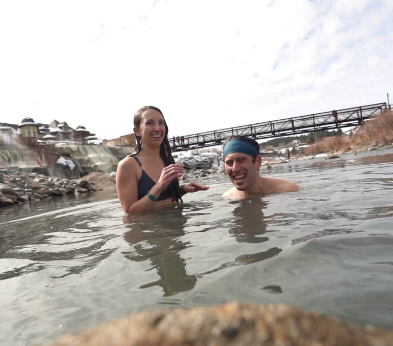 A man and woman laugh, chest-deep in steamy outdoor water, with resort buildings and a distant bridge in the snowy background.