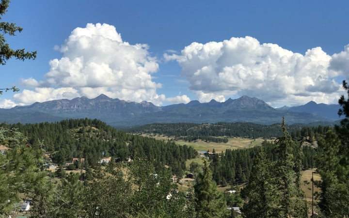 Panoramic mountain range stretches under a blue sky with large cumulus clouds. Coniferous forests cover hillsides with a valley containing scattered homes in the foreground.