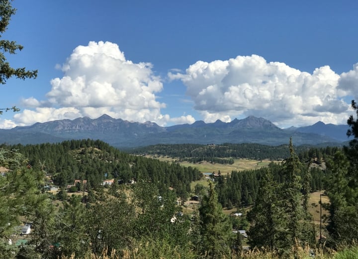 Panoramic mountain range stretches under a blue sky with large cumulus clouds. Coniferous forests cover hillsides with a valley containing scattered homes in the foreground.