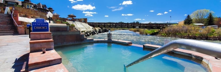 A turquoise outdoor hot spring pool with steps and a handrail sits beside a flowing river and wooden bridge, under a blue, cloudy sky. 
