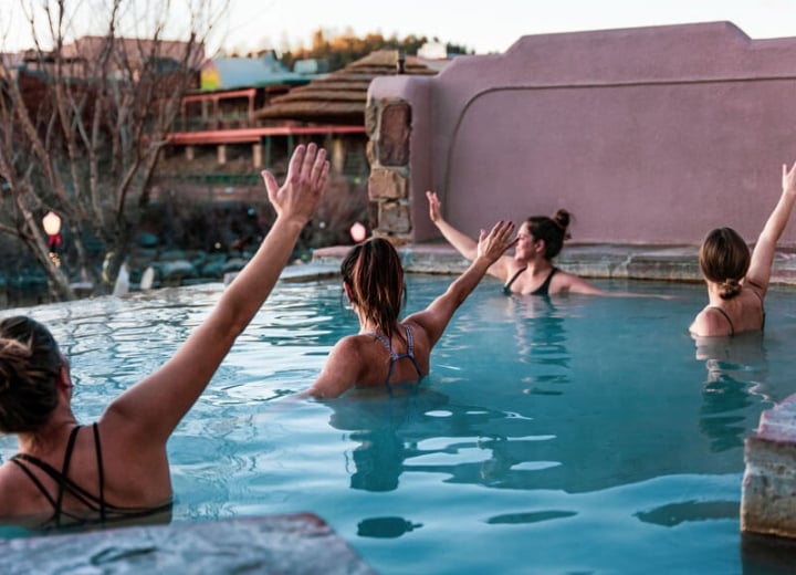 Four women with arms raised are in an outdoor light blue hot spring pool, flanked by reddish adobe walls and distant buildings.