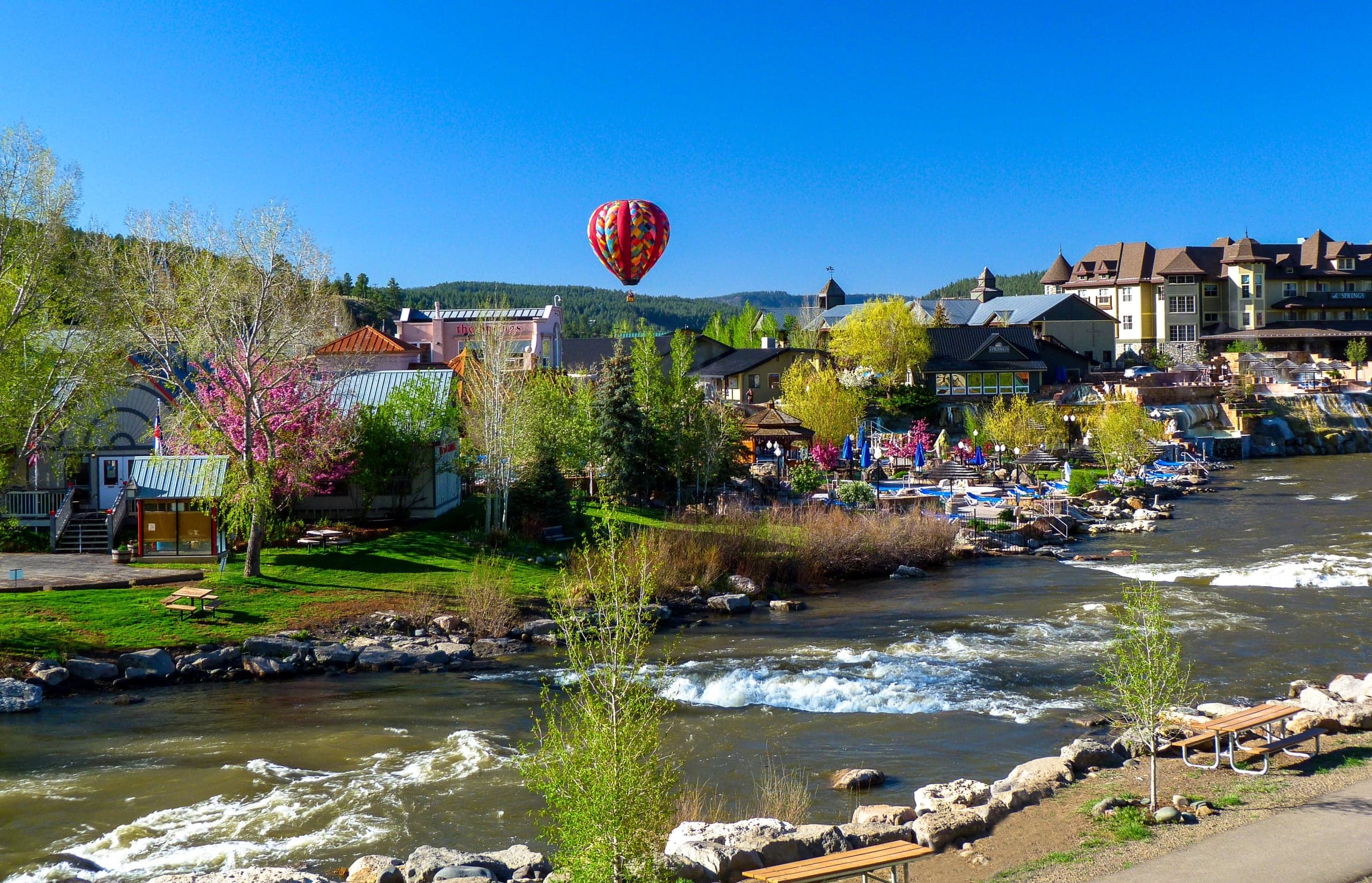 A vibrant hot air balloon floats above a river town. Buildings like 