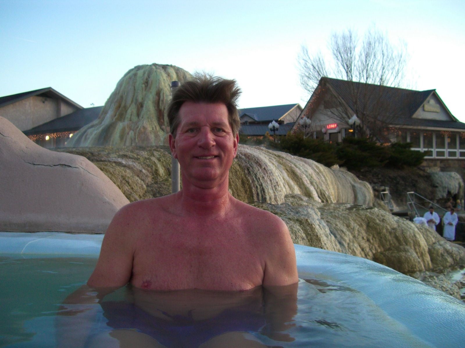 A smiling, shirtless man is partially submerged in water, in a hot spring with natural rock formations and distant buildings, one marked 