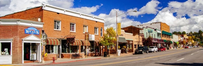 Brick storefronts line a street with parked cars under a bright blue sky with white clouds. Visible text: 