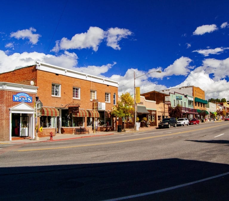 Brick storefronts line a street with parked cars under a bright blue sky with white clouds. Visible text: 