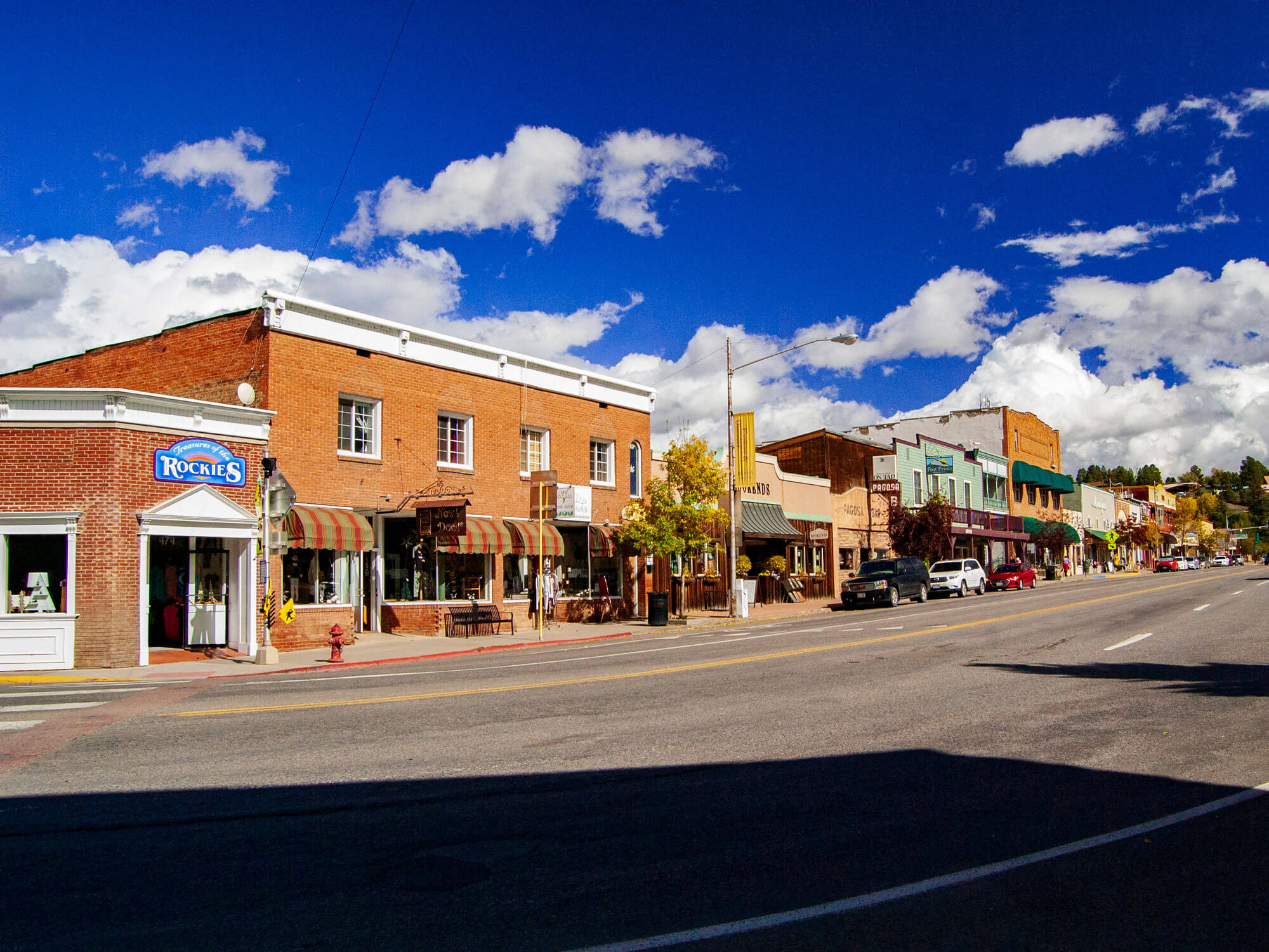 Brick storefronts line a street with parked cars under a bright blue sky with white clouds. Visible text: 