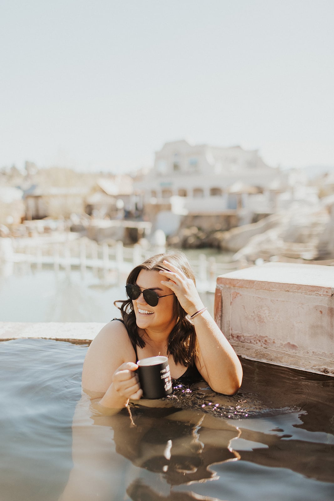 A smiling woman in sunglasses soaks in a hot spring, holding a black mug that reads 'POUR ME SOME SOUL IN A CUP'. Blurred buildings and water are in the bright background.