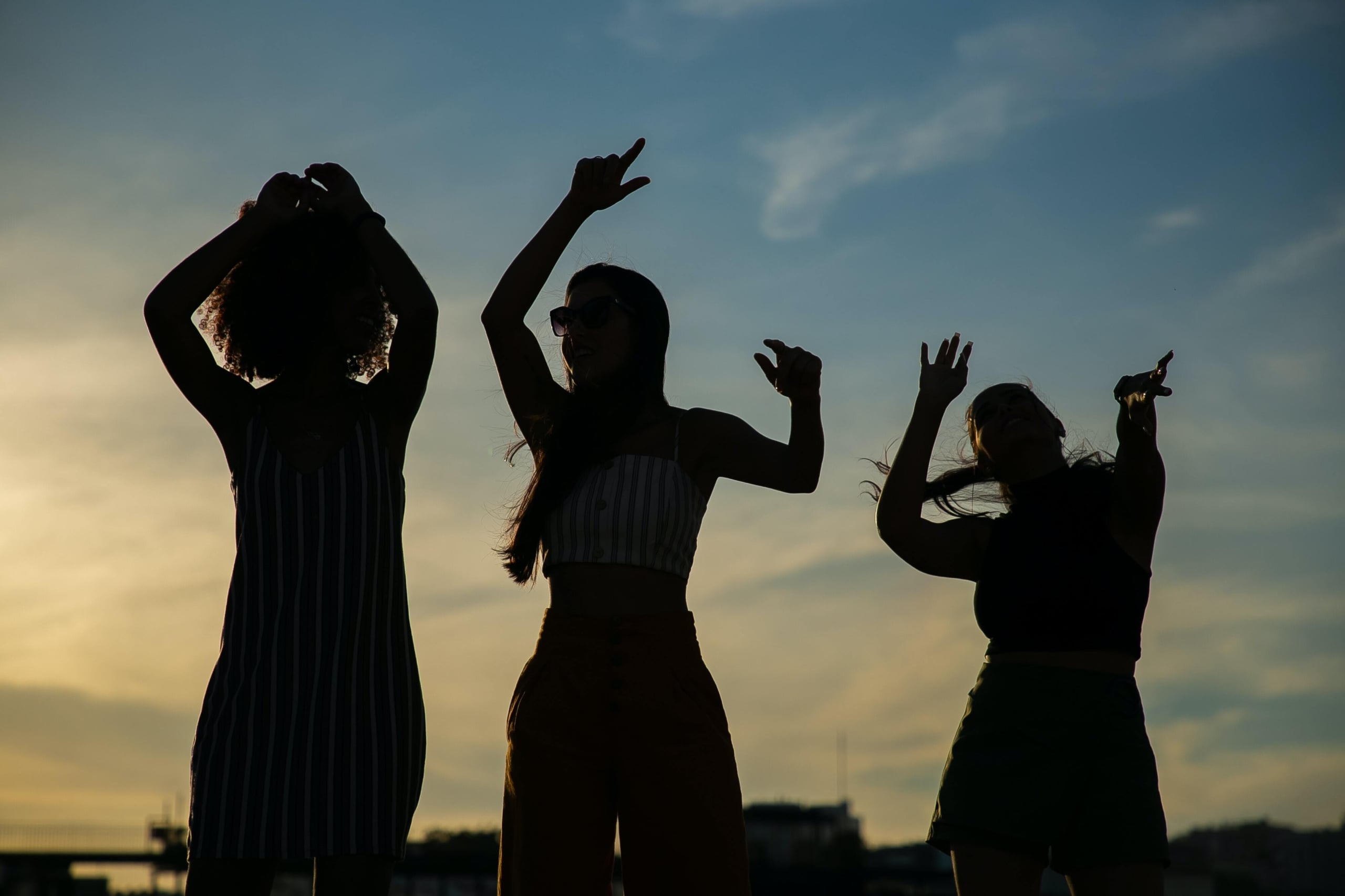 Three silhouetted young women dance with raised arms against a vivid sunset sky, with a city skyline barely visible below.