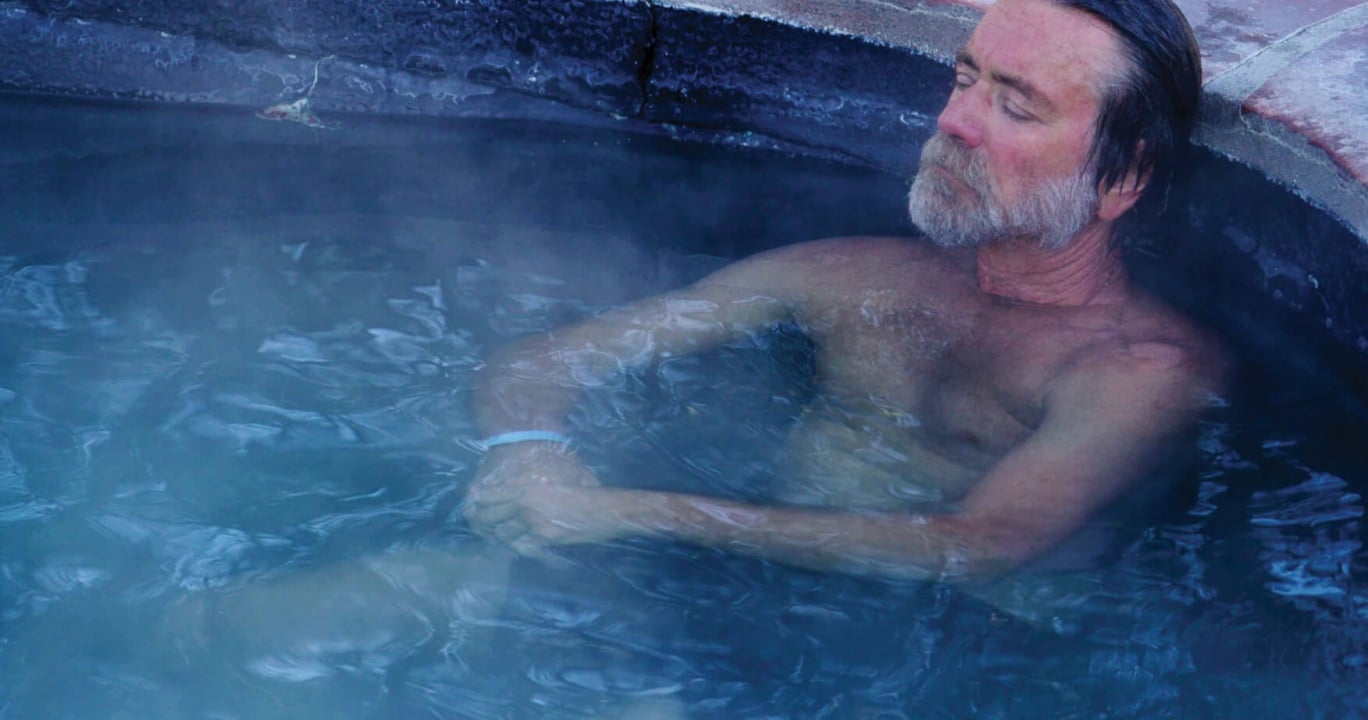 A grey-bearded man reclines with closed eyes, chest-deep in steaming blue water within a rustic red-edged hot tub.