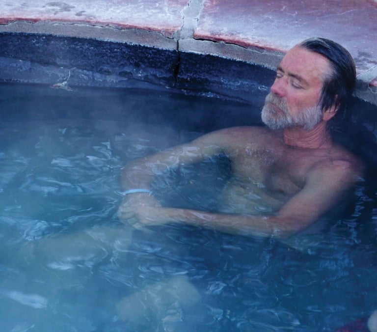 A grey-bearded man reclines with closed eyes, chest-deep in steaming blue water within a rustic red-edged hot tub.