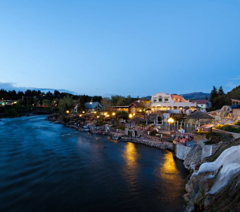 Illuminated hot spring pools with people soaking cascade down a rock formation next to a wide, dark river at dusk, with buildings and hills beyond.