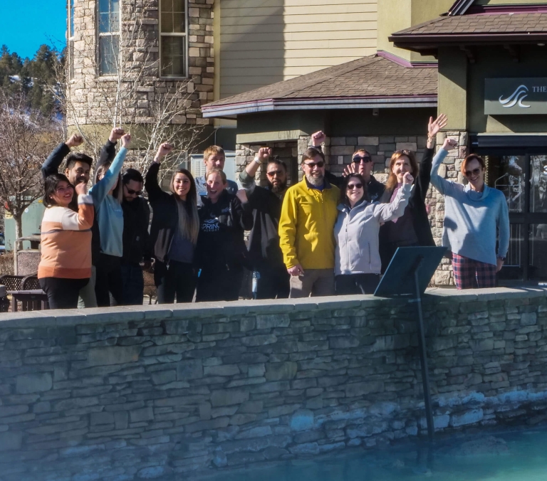 Fourteen diverse people stand on a stone wall, some raising fists, others waving, in front of a multi-story building. A pool of blue water is below. Sign text: THE