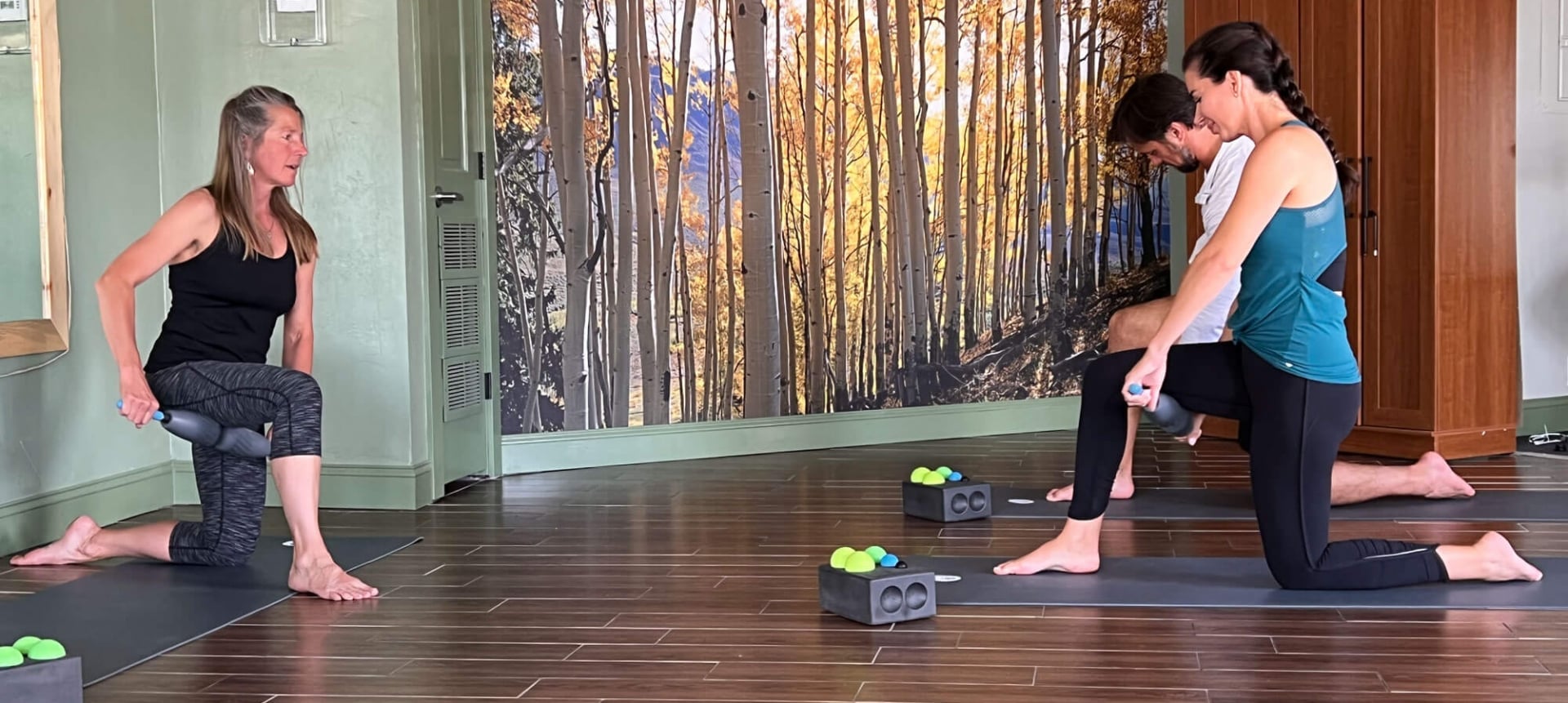 Three people kneel on mats, using rollers for exercise in a studio with green walls and a large mural depicting golden aspen trees.
