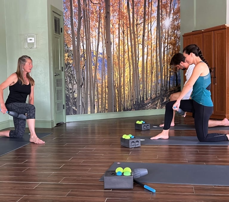 Three people kneel on mats, using rollers for exercise in a studio with green walls and a large mural depicting golden aspen trees.