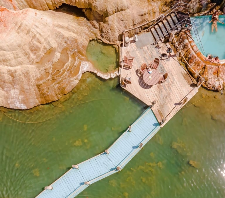 People relax in a blue-water hot tub, accessed by a wooden deck with seating and a boardwalk, amidst layered tan rock formations and green-tinted water.