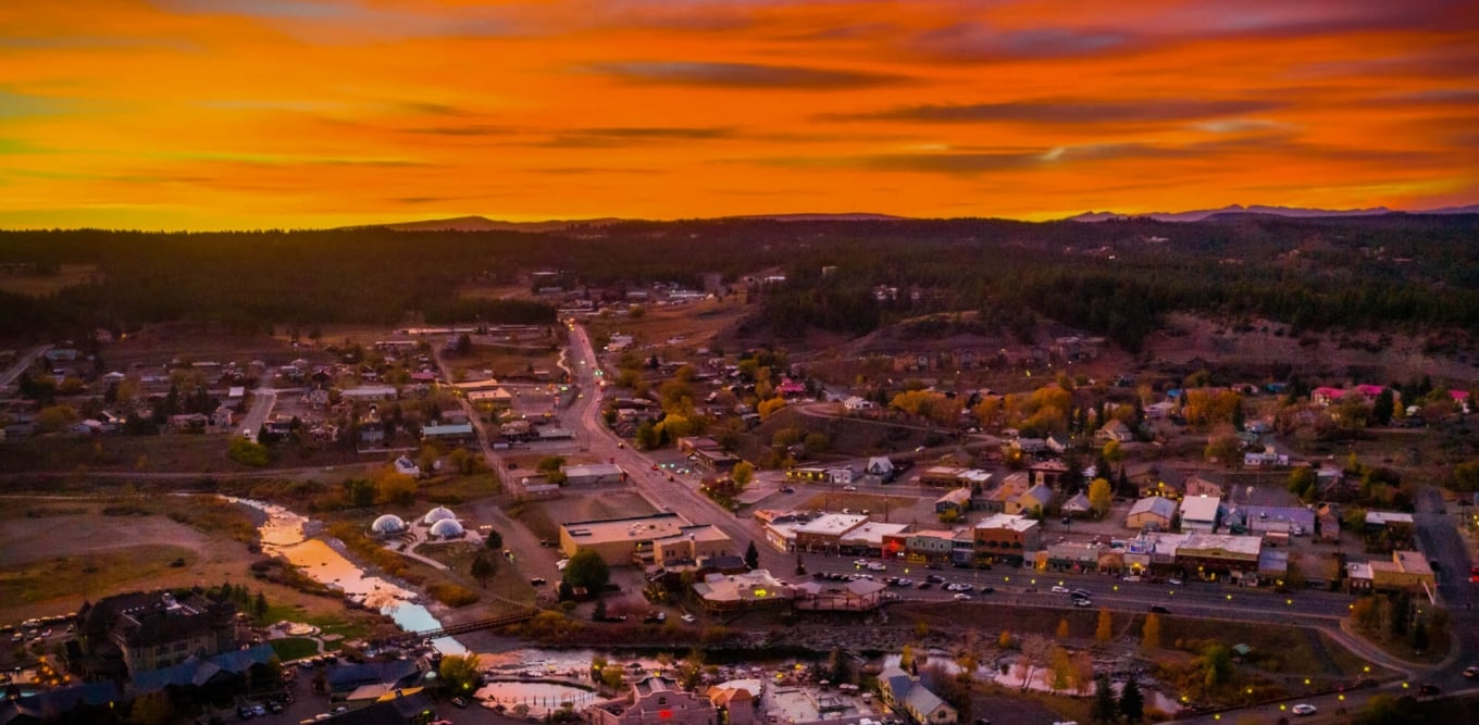 An aerial view of a small town, lights illuminating streets and buildings, a river flowing through, all set amidst forested hills under a fiery orange sunset.
