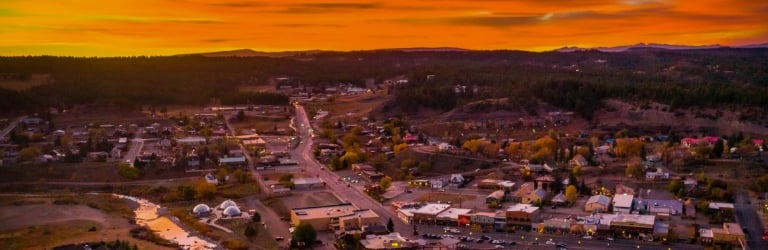 An aerial view of a small town, lights illuminating streets and buildings, a river flowing through, all set amidst forested hills under a fiery orange sunset.