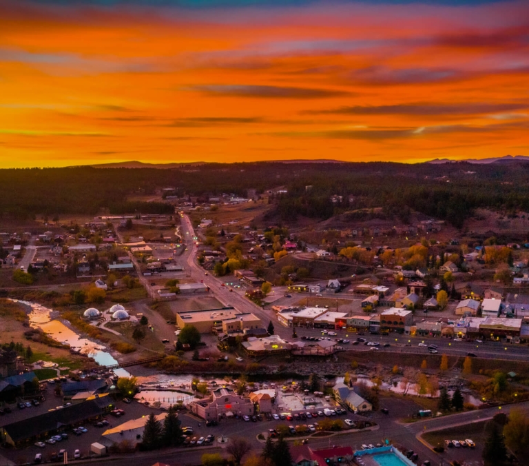 An aerial view of a small town, lights illuminating streets and buildings, a river flowing through, all set amidst forested hills under a fiery orange sunset.