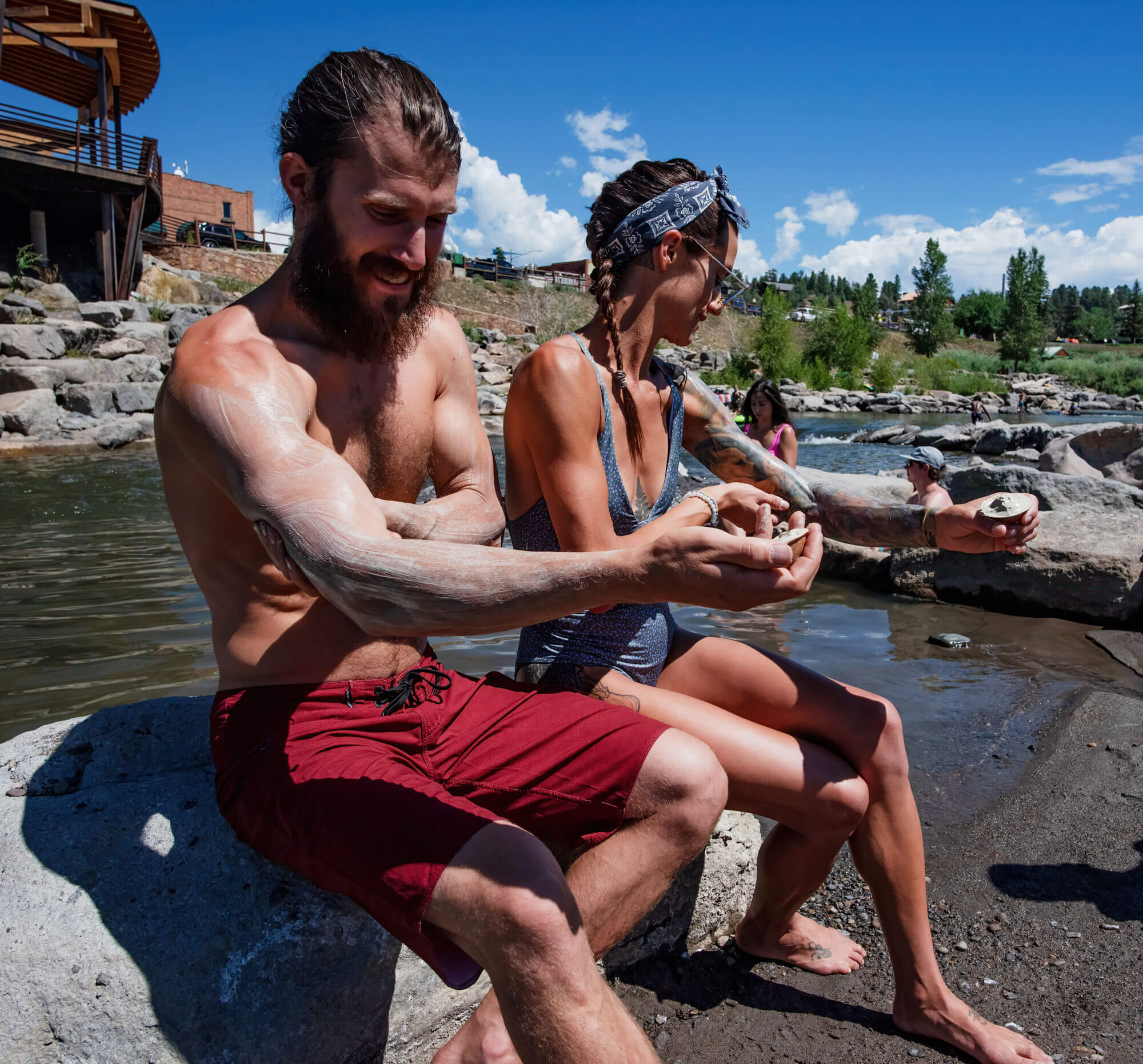 A man and woman apply mud/clay to their bodies using shells, sitting on rocks by a river with others on a sunny day.